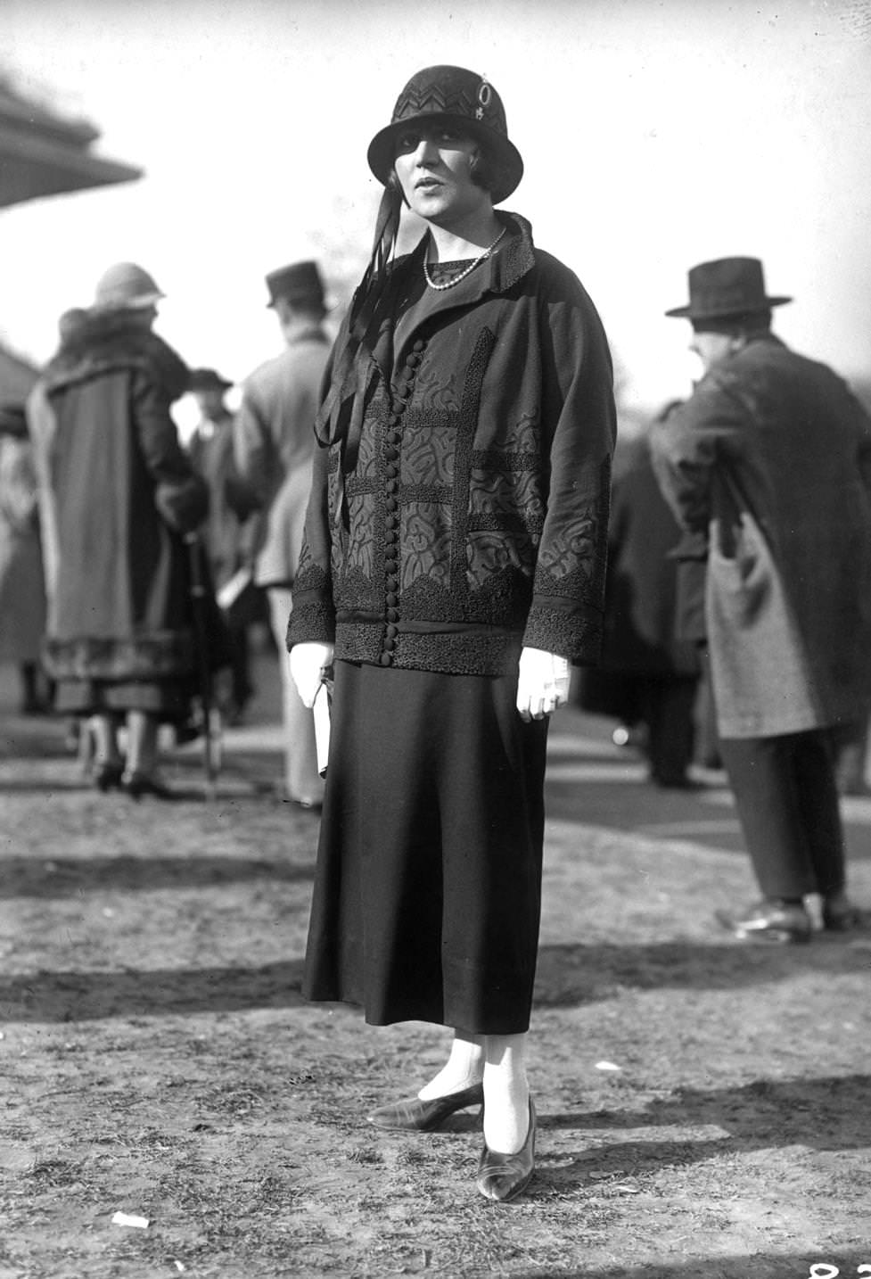 A short embroidered jacket with cloth covered buttons worn over a plain skirt. A cloche hat is worn with a decoration of long ribbons hanging down under the brim, 1924