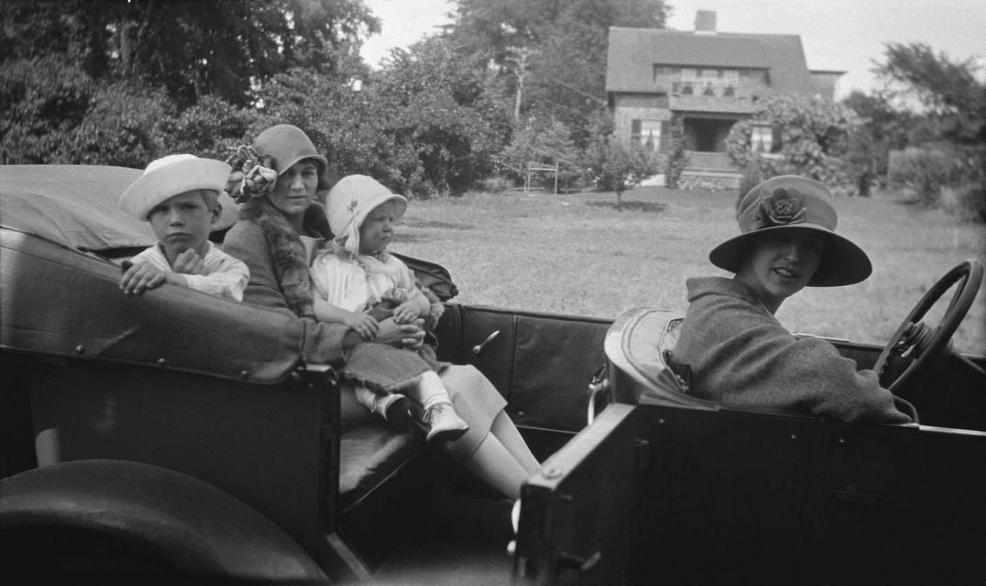 Two elegantly dressed women, one in a cloche hat the other in a wide brimmed hat, and two smartly dressed children all sitting in car outside a large gardened house, 1924.