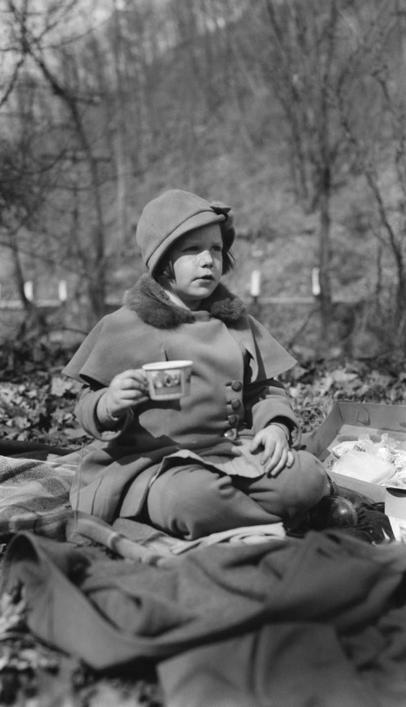 Young, smartly dressed child (girl), wearing caped coat and cloche type hat, sitting on blankets outdoors having a picnic, 1924.