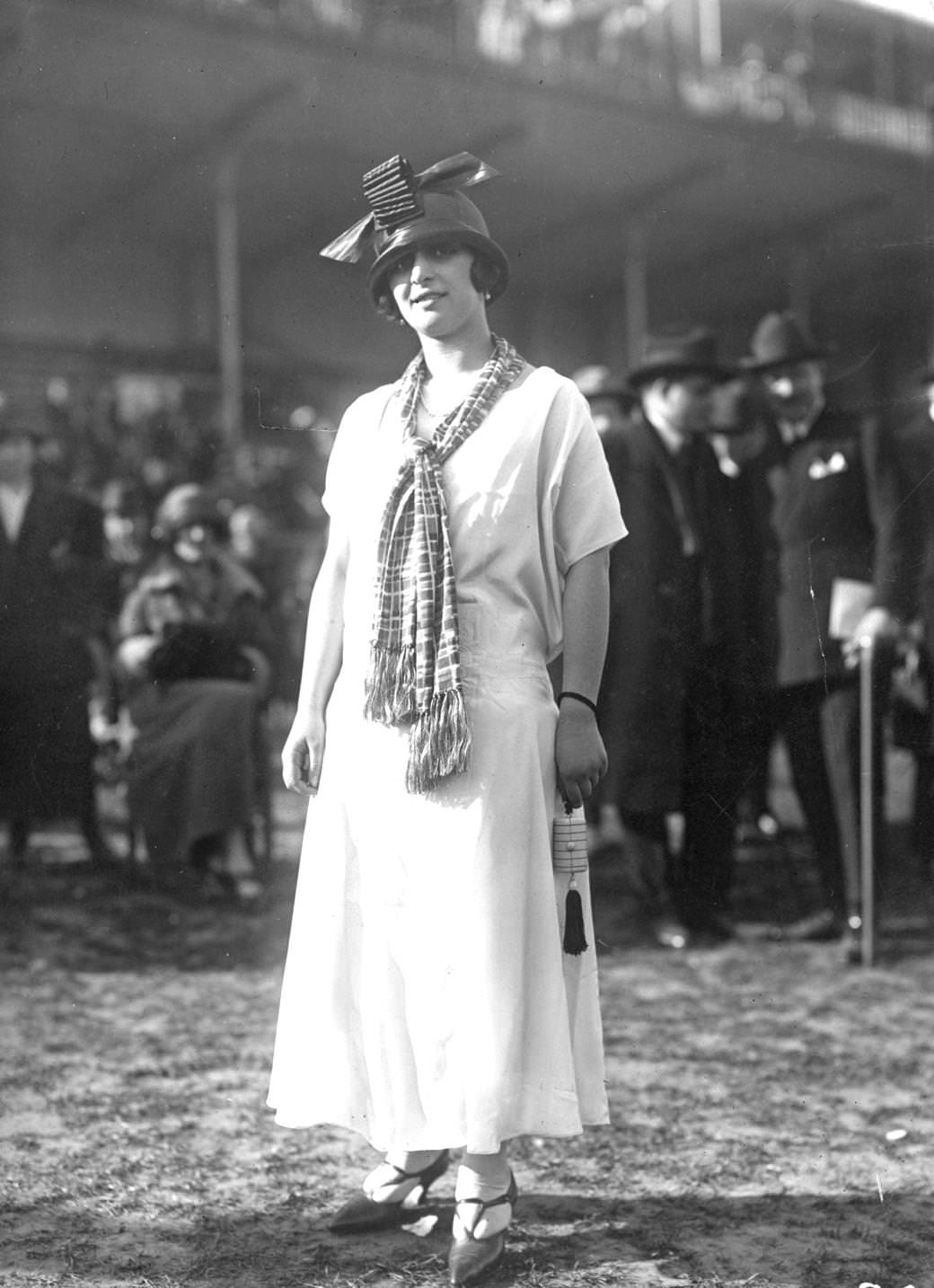 Simple, loose waisted short-sleeved dress worn with a loosely knotted scarf. A hat is cloche shaped with a large bow on the front, 1924
