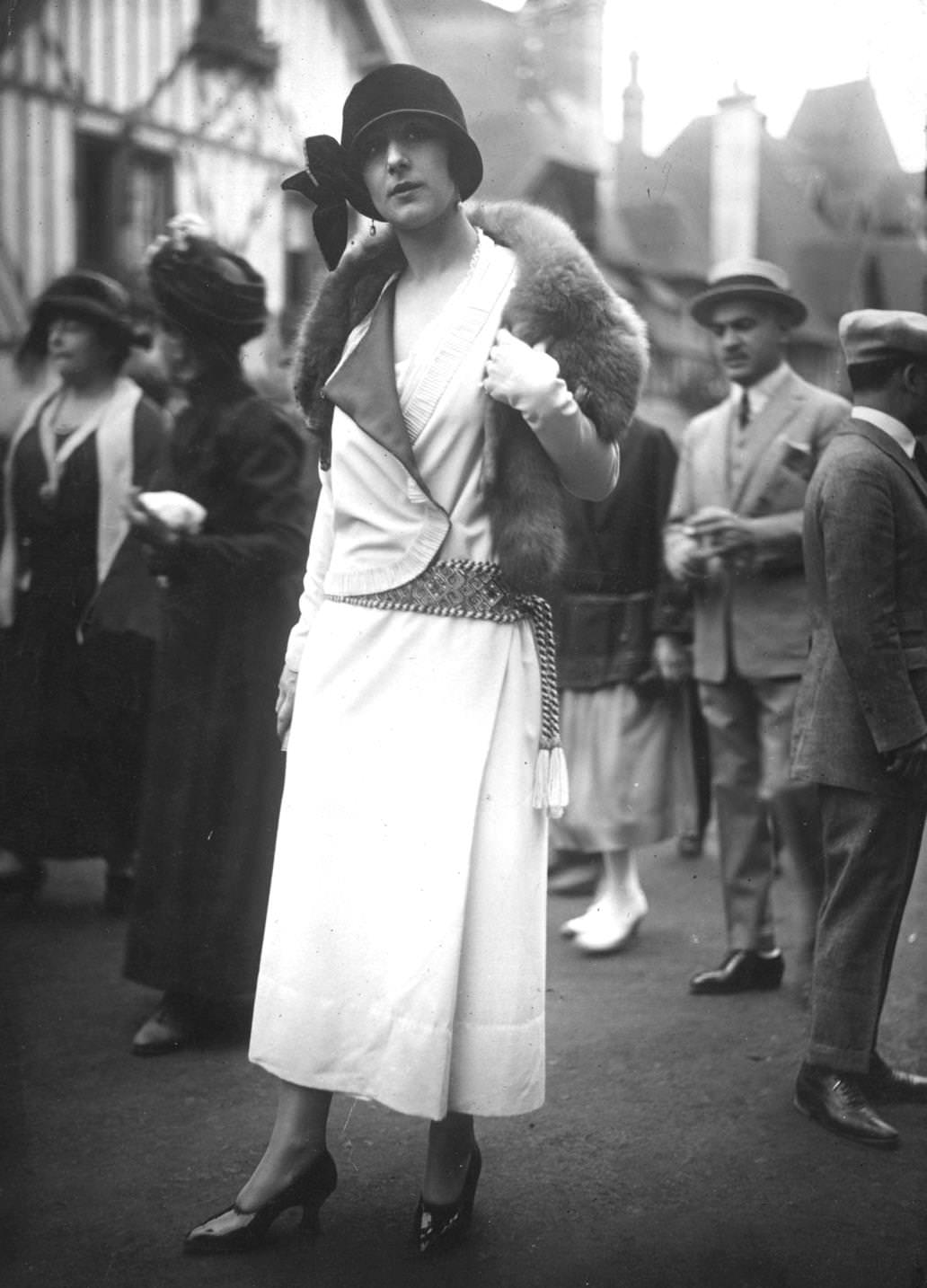 The model wears a wrap over coat with a low-waisted embroidered and tasselled belt, with a fur stole and a cloche hat decorated with a velvet bow, 1923
