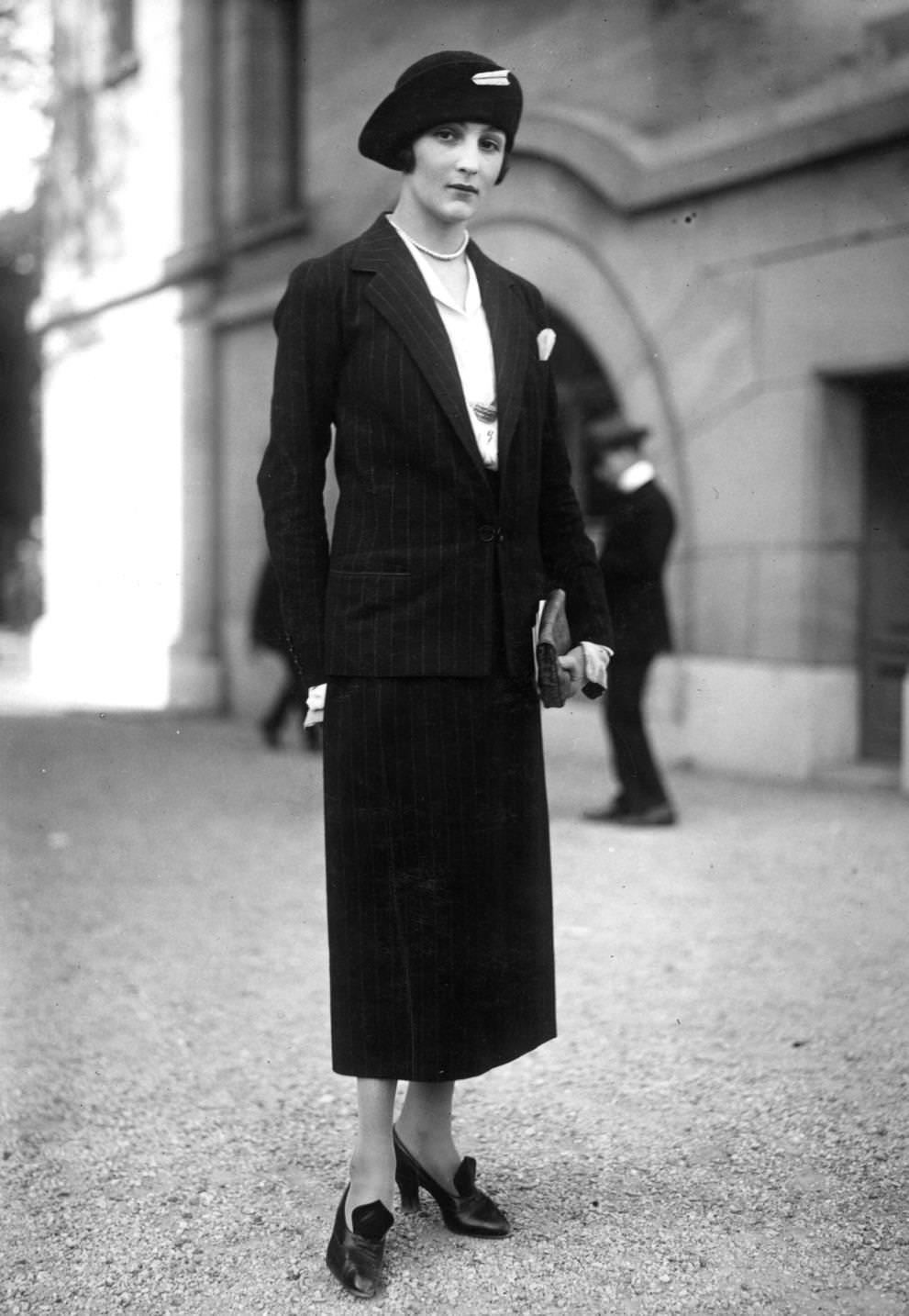 Pin striped suit with a slim skirt. Cloche hat has a rolled back brim with a decorative hat pin. Jewellery is a short single strand bead necklace, 1923