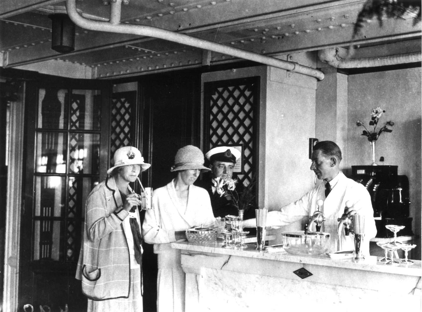 A barman serves drinks to a couple of women at the soda fountain on board the Cunard liner Aquitania, 1920s