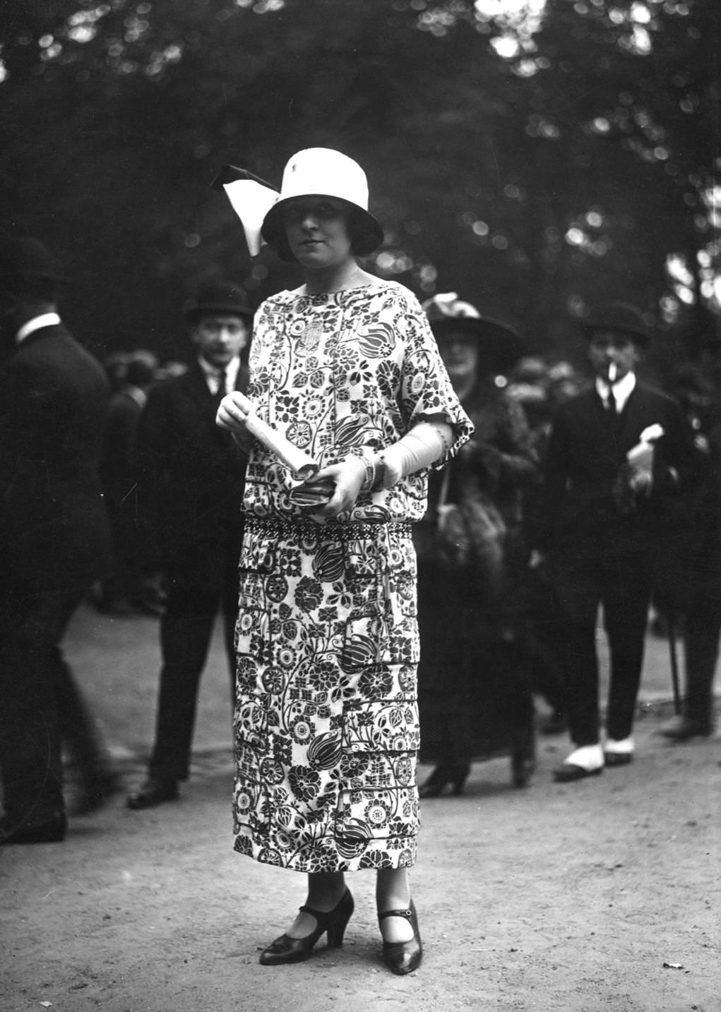 Drop-waisted ankle length dress in multi-patterned material with three-quarter sleeves and elbow length gloves. Worn with a cloche hat decorated with a bow. One-bar shoes, 1923