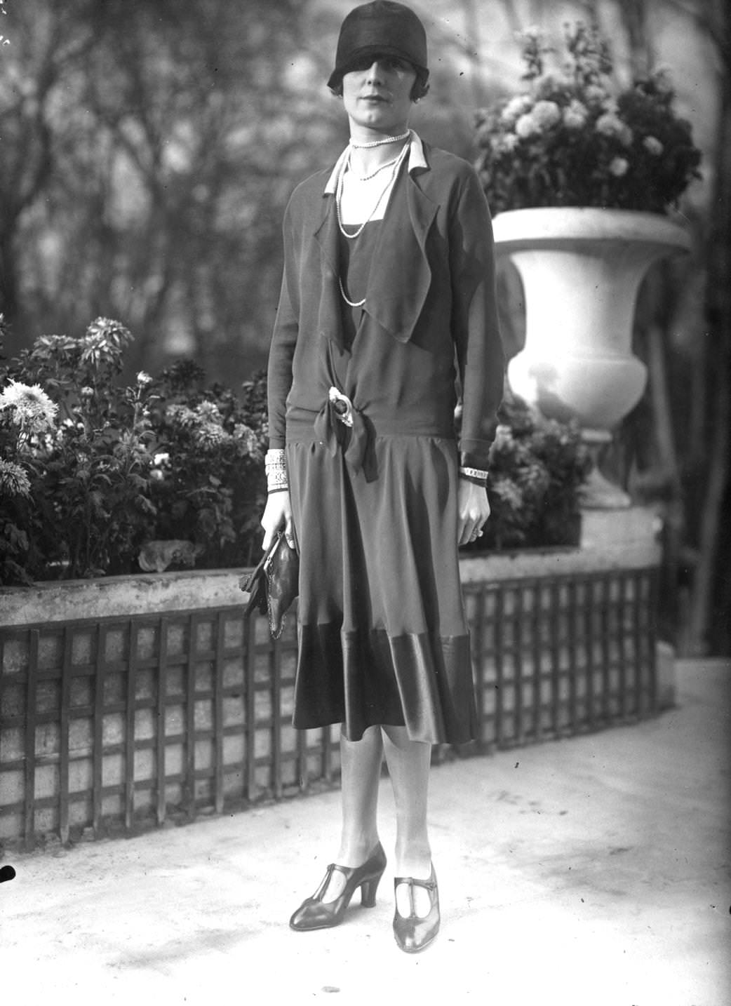 Long waisted dress with a tie on the hips, v-necked with a deep satin edge to the skirt. Accessories are a cloche hat, ropes of beads and several bracelets worn over long sleeves, 1925