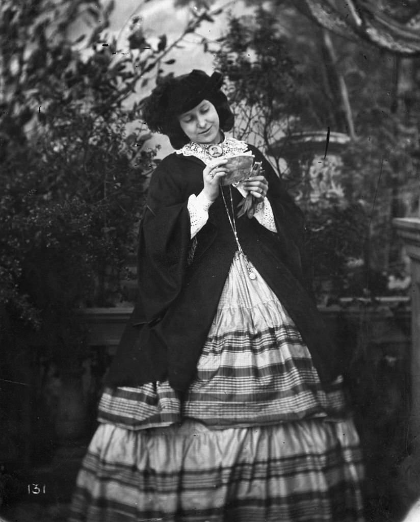A woman wearing a crinoline, typical dress of the mid 1800's, reads a letter in a garden setting, 1860