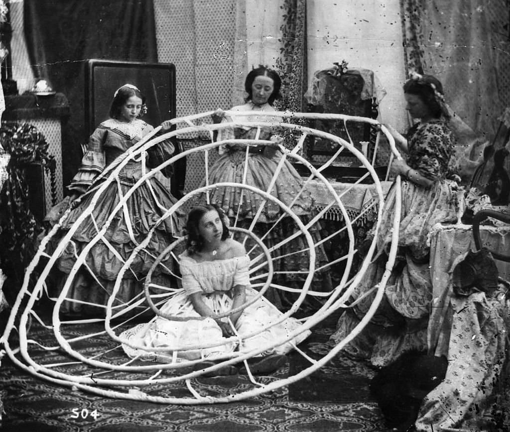 A woman wearing a crinoline being dressed with the aid of long poles to lift her dress over the hoops.