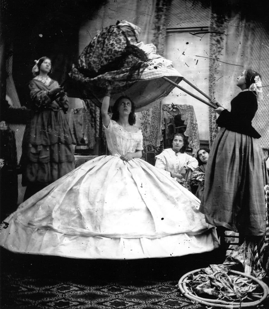 A woman wearing a crinoline being dressed with the aid of long poles to lift her dress over the hoops.