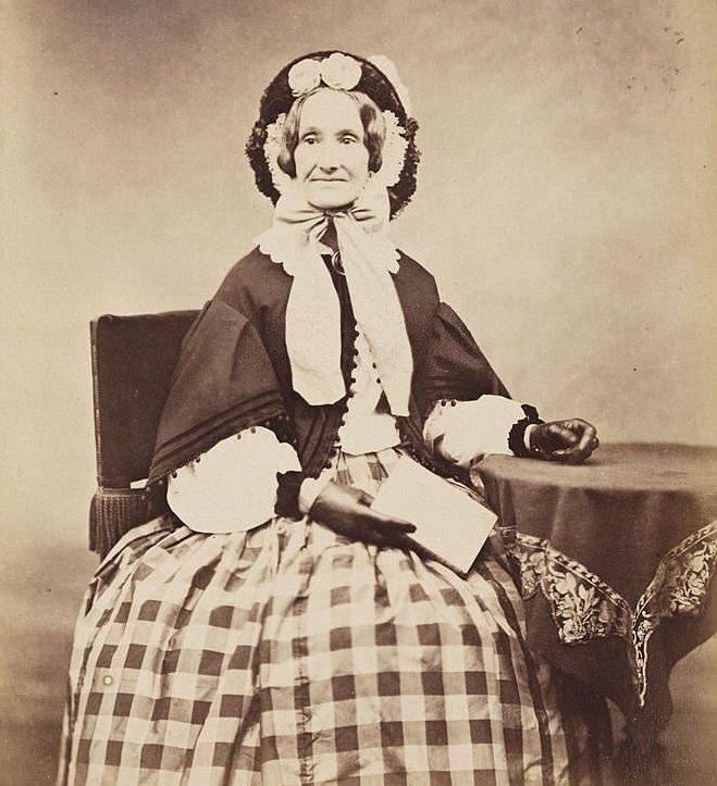 Victorian woman in a bonnet and checked skirt, with a book in one hand, 1860