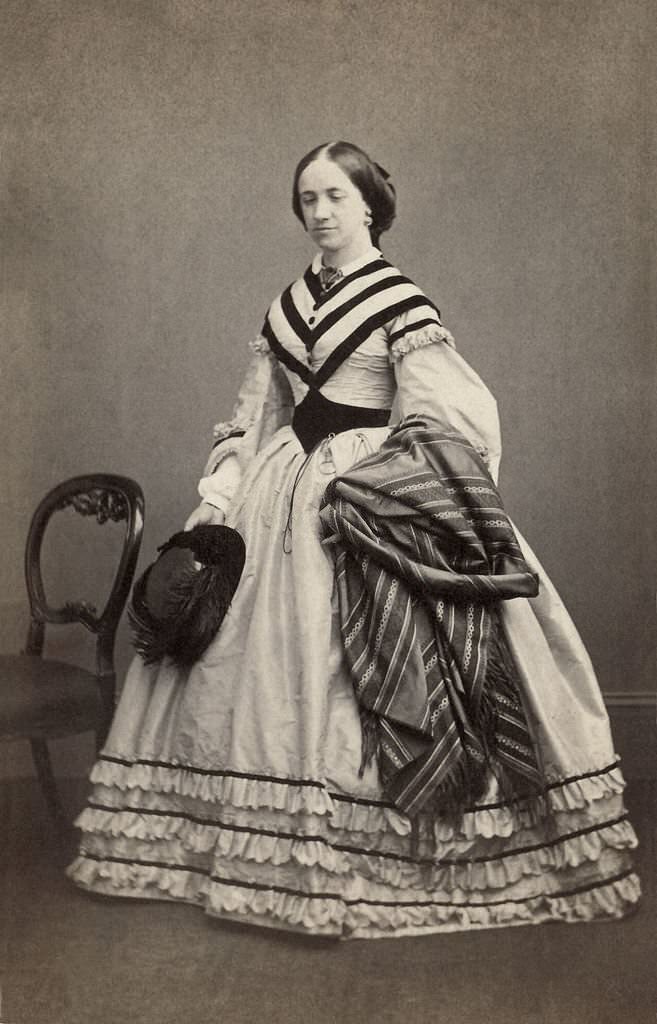 The woman carries a feathered hat and a piece of striped fabric, possibly a shawl. She wears a monocle or pince-nez at her waist, 1856