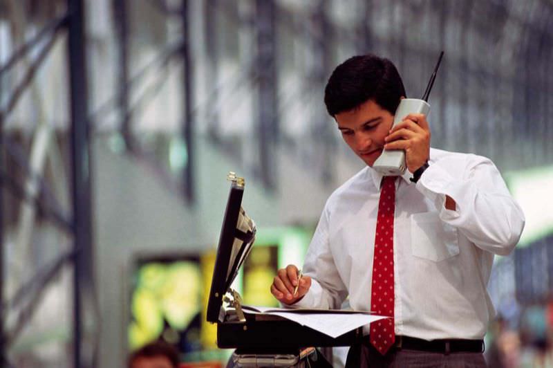 A young businessman with a brief case goes over figures on a giant old cell phone, 1990.