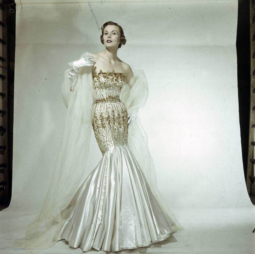 A model presenting for Jacques Fath, a gold-embroidered satin sheath dress, which flares out in godets at the bottom.