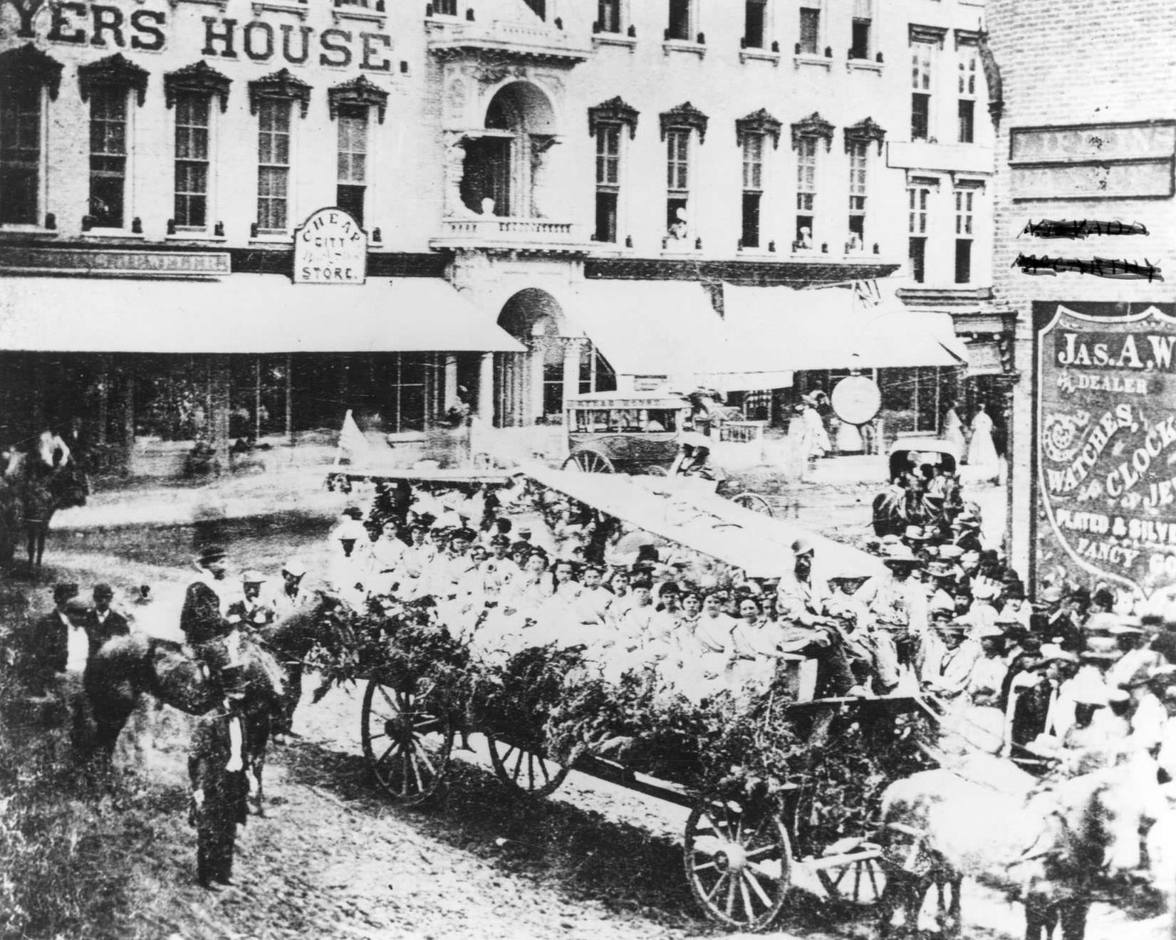 A horse-drawn float full of flower-bedecked women dressed in white riding in an 1869 July 4th parade in front of the Myers House, 1869