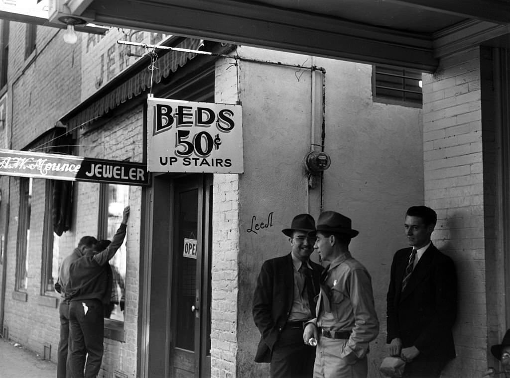 Workers standing outside of a housing facility where the sign reads 'Beds 50 cents up stairs', December 1940