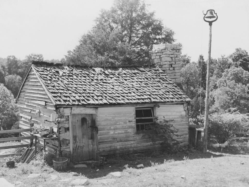 A small wooden shack, with a shingled roof, brick chimney stack, and a bell set on top of a tall pole at right, Kentucky, 1940