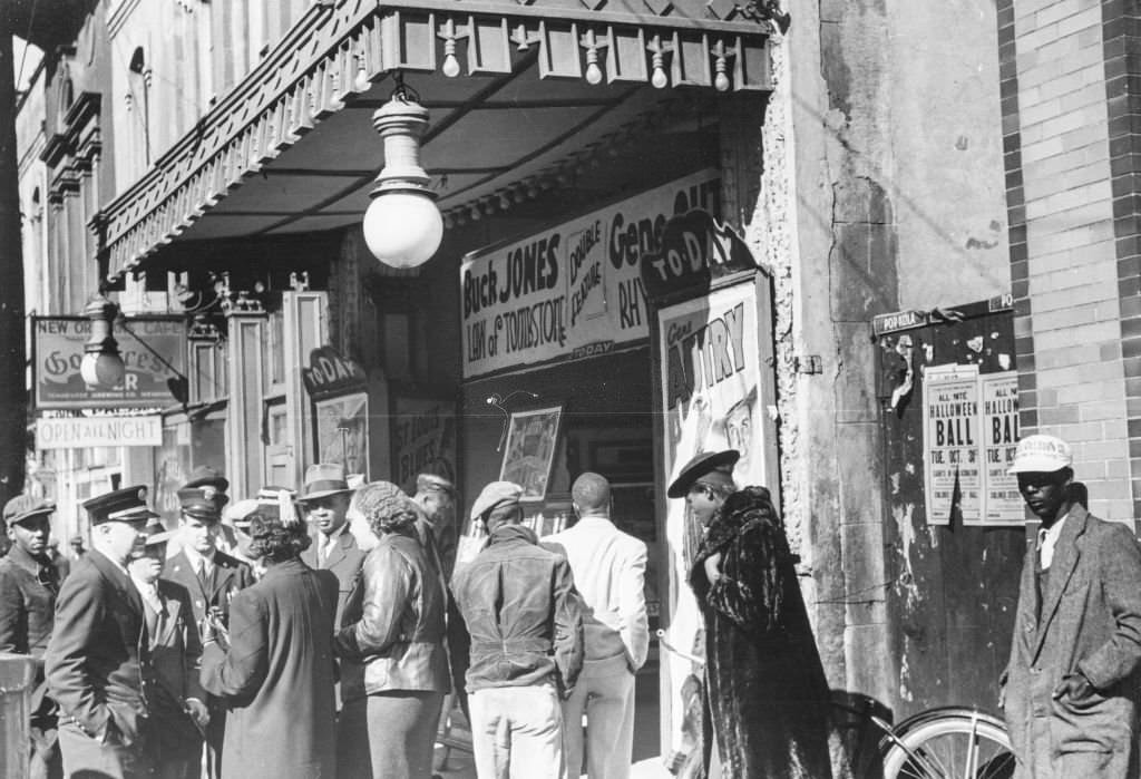 A group of men and women standing outside a cinema waiting to be allowed in, with signs advertising "Buck Jones, Law of Tombstone, " and "Gene Autry"; located on Beale Street in Memphis, 1939