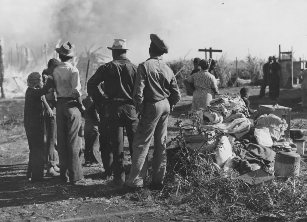 A small group of African American men, women, and children, standing together, with their backs to the camera, watching a fire in the left background, Florida, 1939