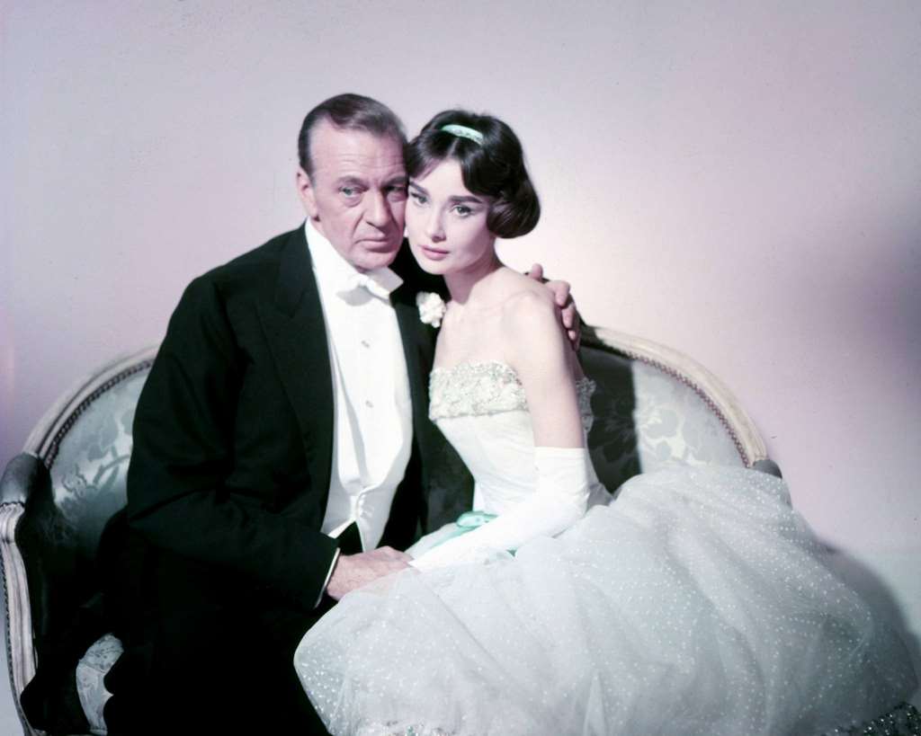 Gary Cooper and Audrey Hepburn in formal wear, sit on a loveseat in a scene from 'Love in the Afternoon', 1957.