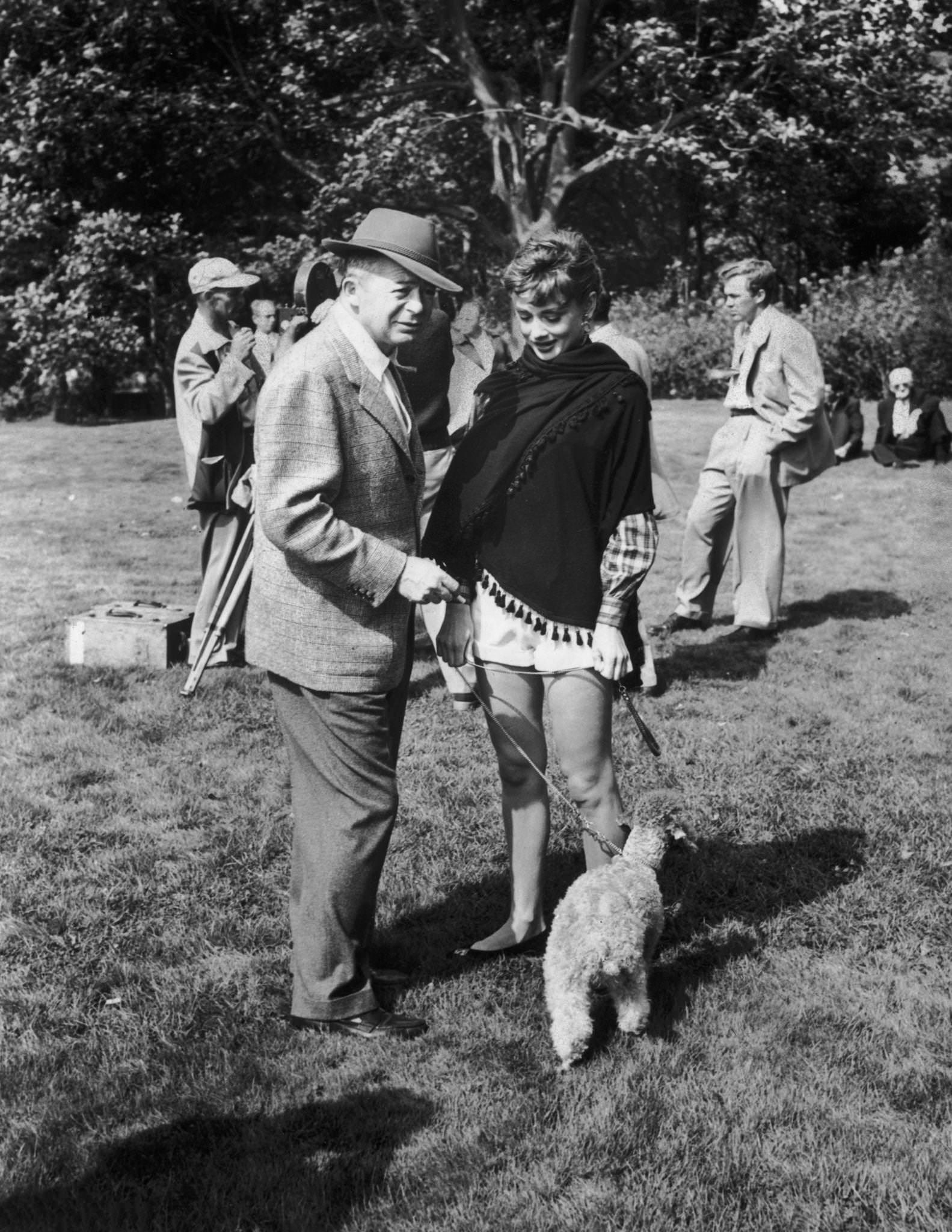 Director Billy Wilder standing outdoors beside Belgian-born actor Audrey Hepburn, who holds a poodle on a leash, on location for his film, 'Love in the Afternoon'.