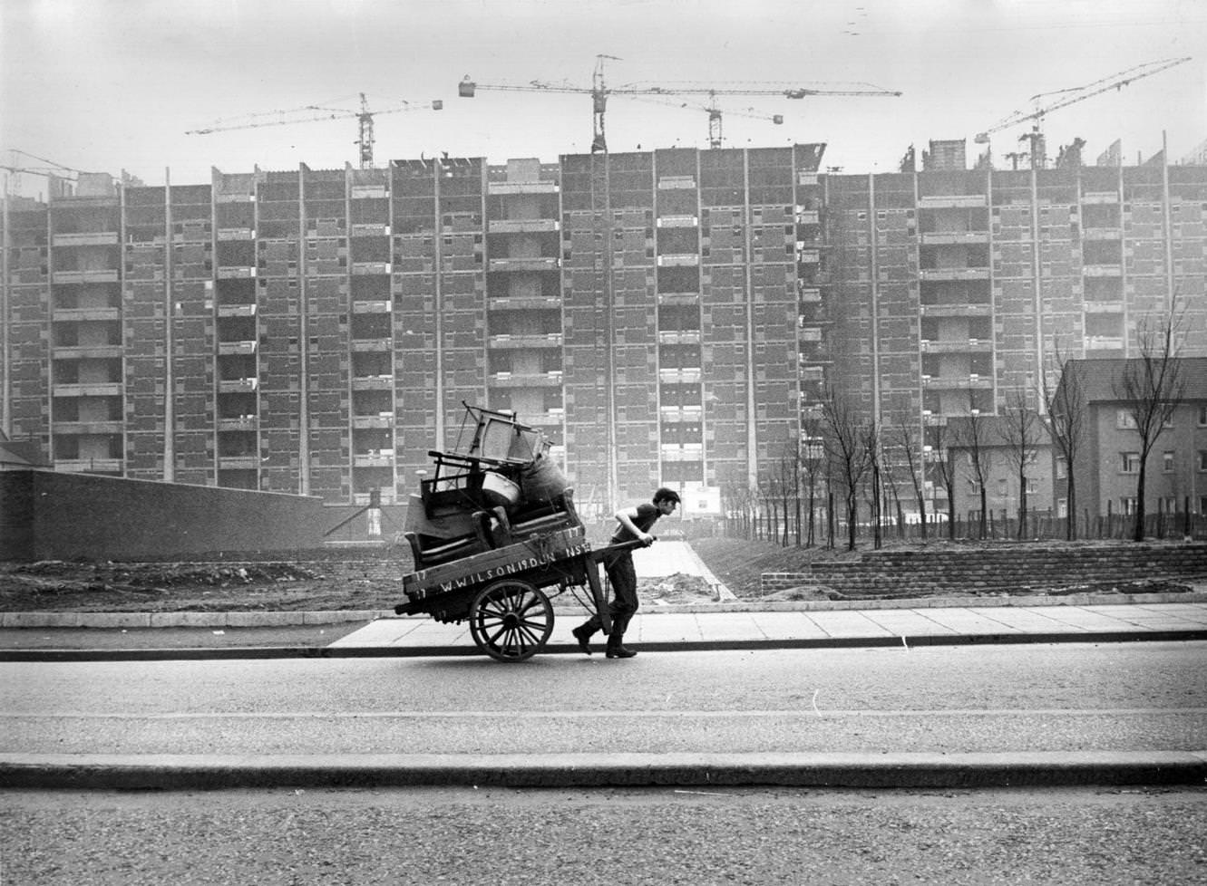 A man pulling a hand cart past tower blocks under construction in the Gorbals area of Glasgow, 1960