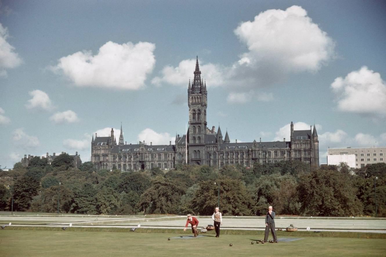 Three men play a game of lawn bowls on a bowling green in West End Park with the New Buildings of the University of Glasgow rising behind on Gilmorehill in Glasgow, Scotland, 1965.