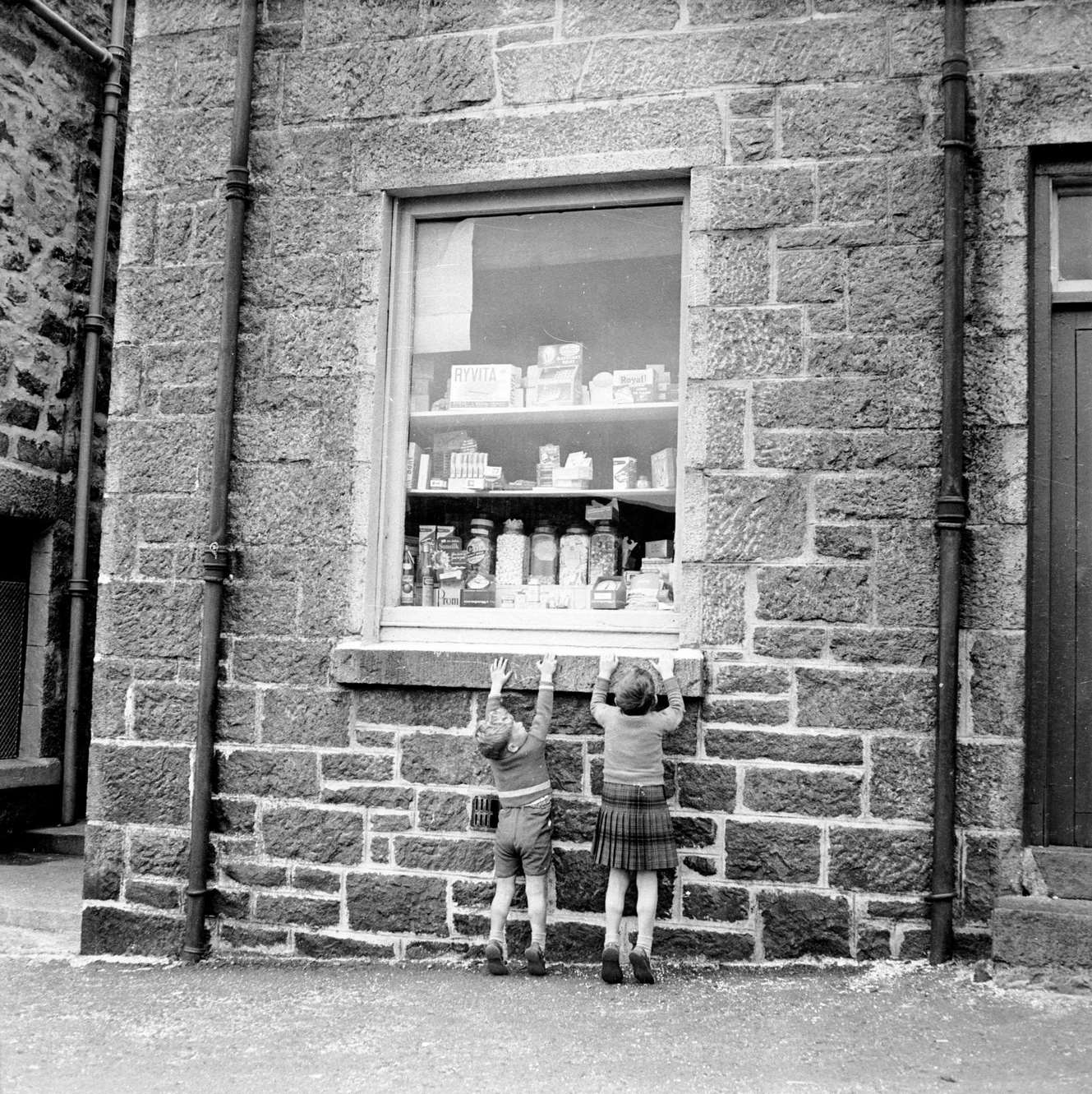 Two children stretching up to see sweets on display in a shop window at Castlebay on the island of Barra in the Outer Hebrides, 1960