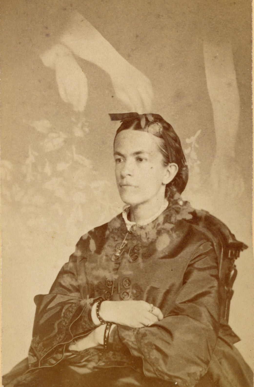 Unidentified young woman seated with her arms crossed. Faint images of three arms and flowers float over her head.