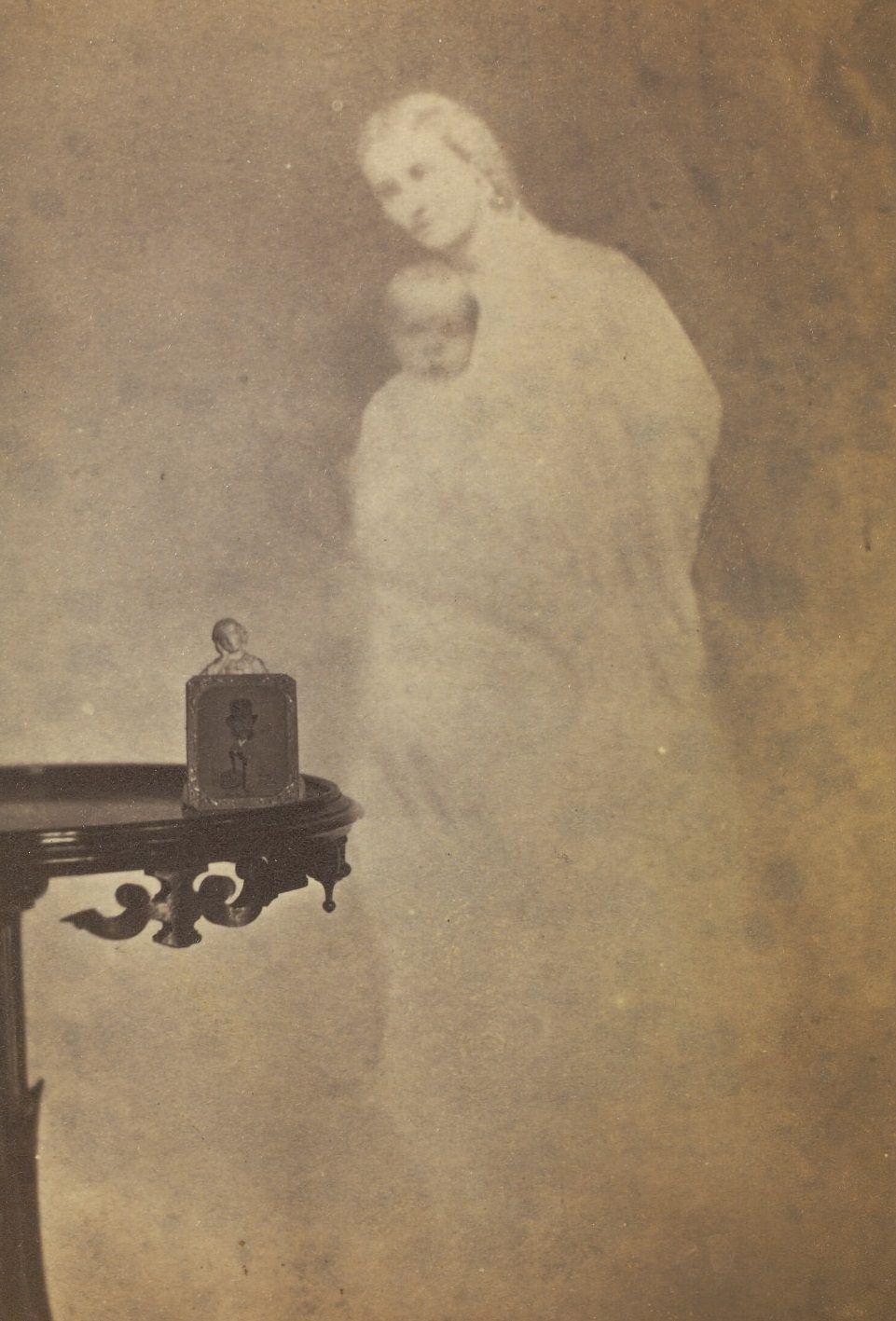 Photograph of a statuette and drawing on a side table, with a faint image of a woman and baby in the background.