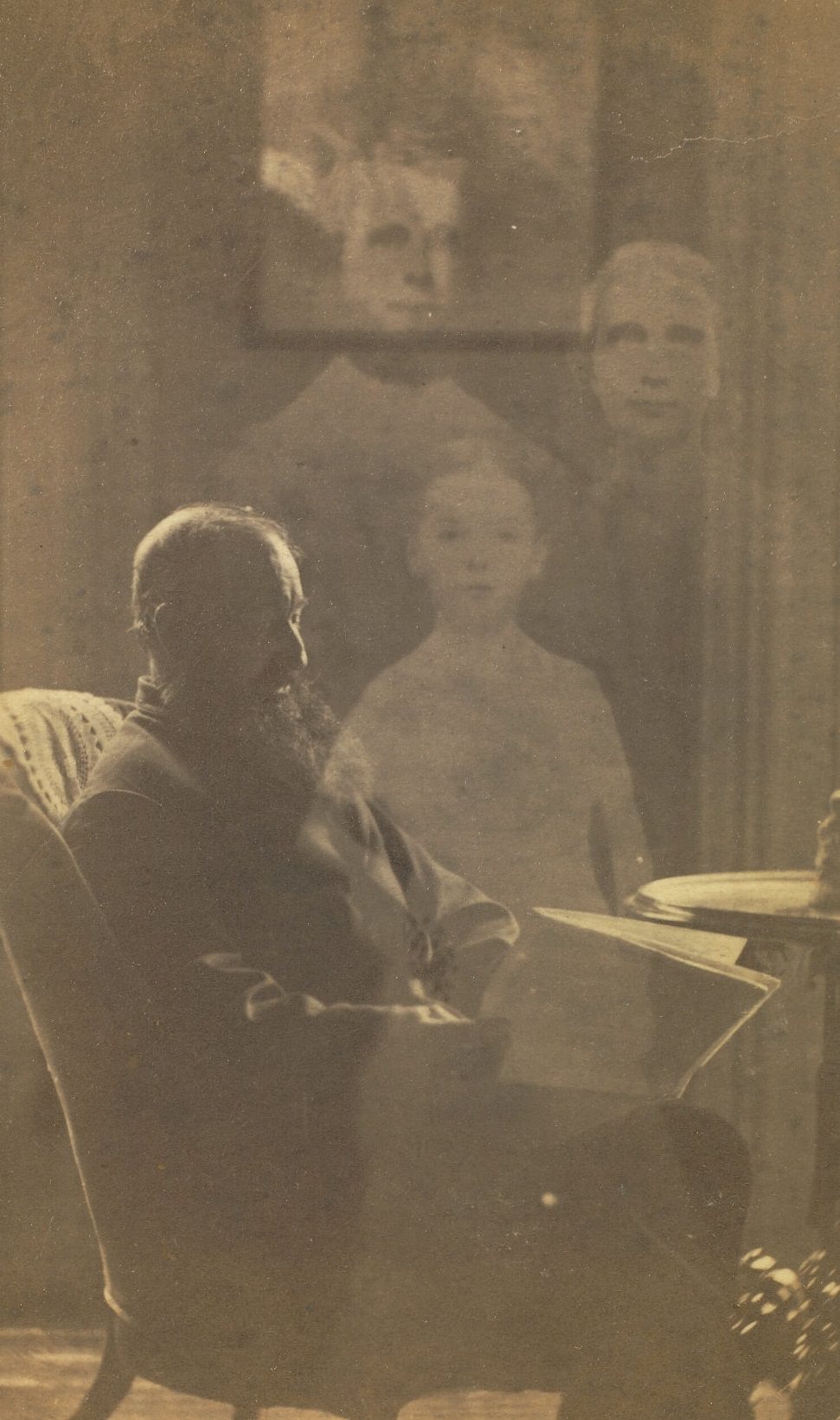 Profile portrait of a man with a beard seated in an upholstered chair, next to a side table. The faint images of three children appear behind him.