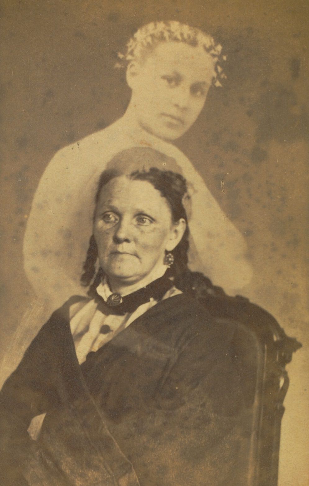 Portrait of a woman seated with her arms crossed. The faint image of a younger woman wearing a floral hair wreath appears above her.