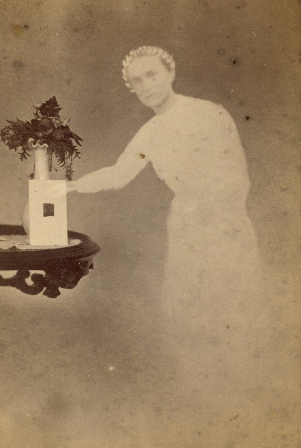 A photograph propped against a vase of flowers on a side table, with the faint image of a woman reaching out with one arm next to the table.