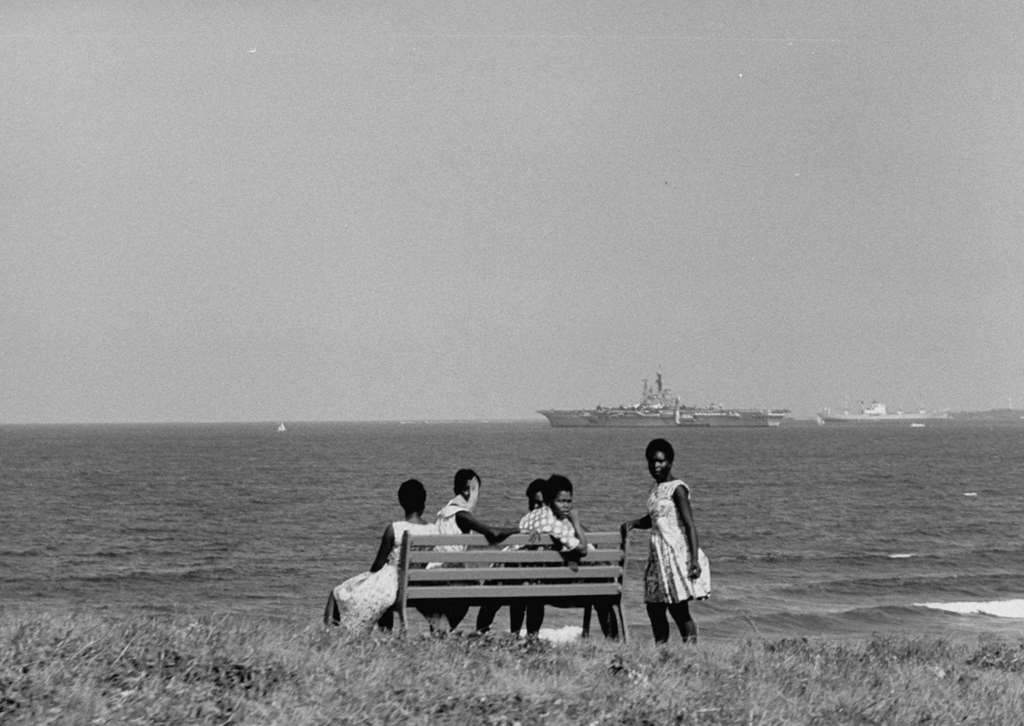 Local residents seated on a bench to watch the Royal Navy light fleet carrier HMS Centaur (R06) on patrol off the coast of Dar es Salaam, 1964