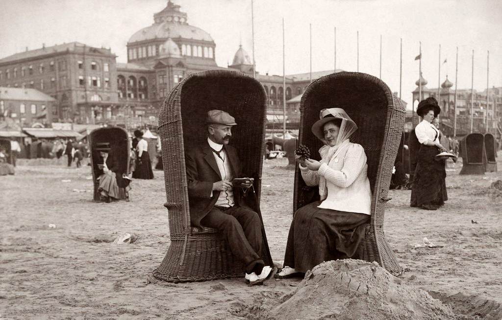 A couple on holiday seated in beach chairs, she eating grapes and he smoking a cigar at the seaside in Scheveningen near The Hague in the Netherlands, 1910.