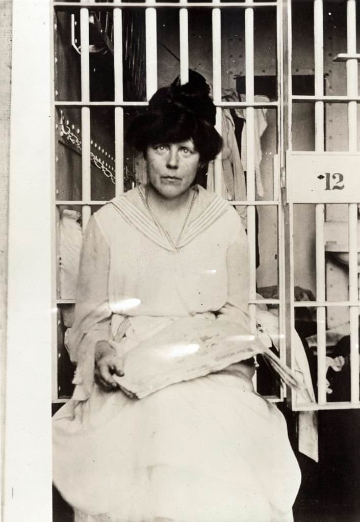 Lucy Burns, three-quarter length, seated, facing forward, holding a newspaper in her lap in front of a prison cell.