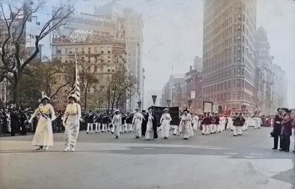 Women wearing white Edwardian clothing, and sashes, and holding a US flag and pennants, followed by a marching band, taking part in a suffrage parade, 1913