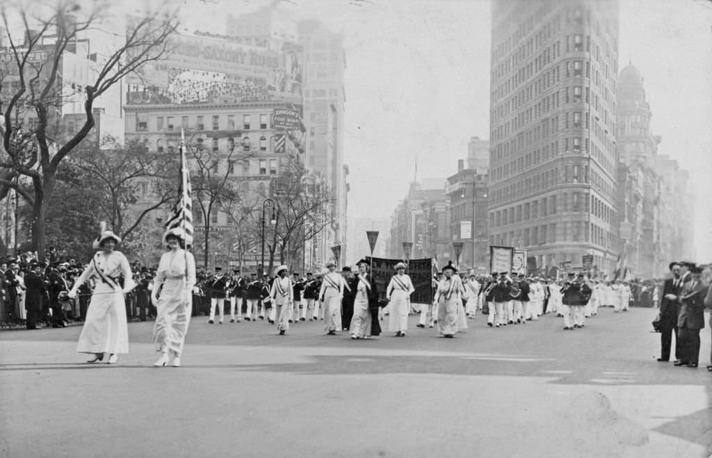 Women wearing white Edwardian clothing, and sashes, and holding a US flag and pennants, followed by a marching band, taking part in a suffrage parade, 1913