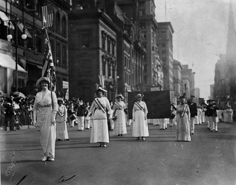 A parade for Women's Suffrage marches down a Manhattan street led by the marshal carrying an American flag, 1913