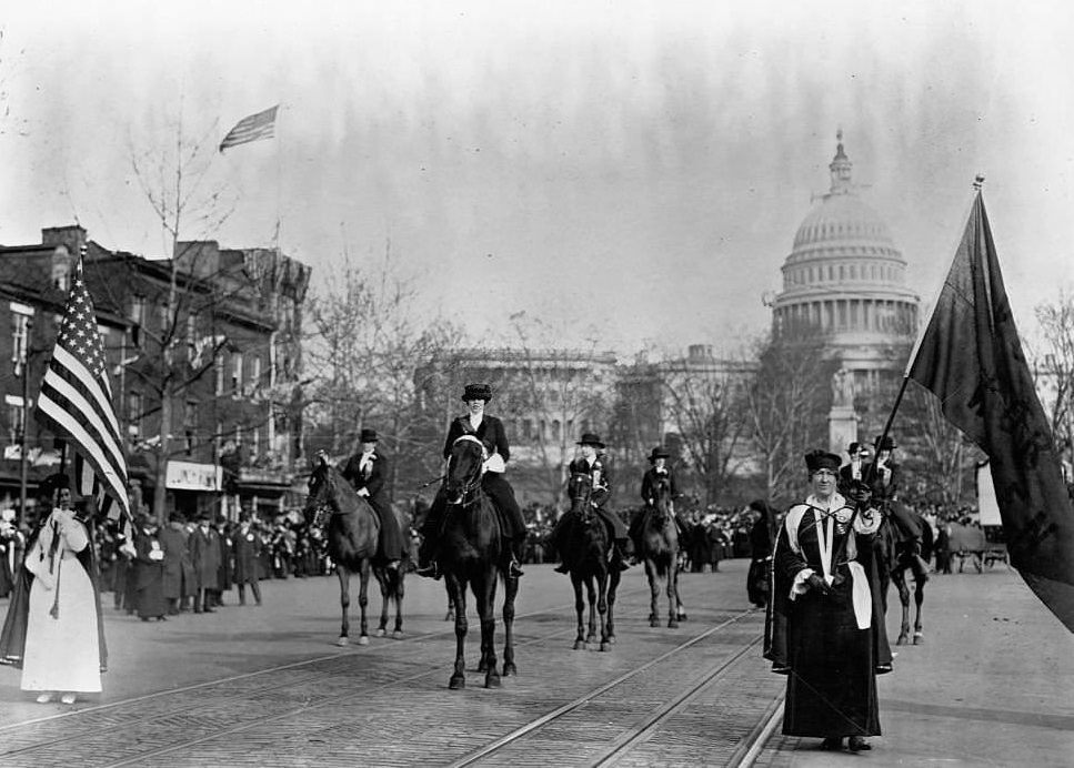 Some women march and carry flags, others ride horses in a suffrage parade on a street by the Capitol Building in 1913. Washington D.C., USA.