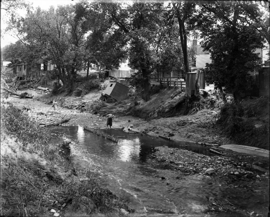 River at low level running through a neighborhood, 1906. People are in the riverbed and a shack that has fallen off its foundation is also visible along the bank.