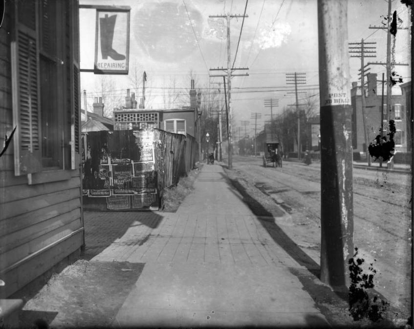 City sidewalk with wooden fence with posters plastered on it, 1907