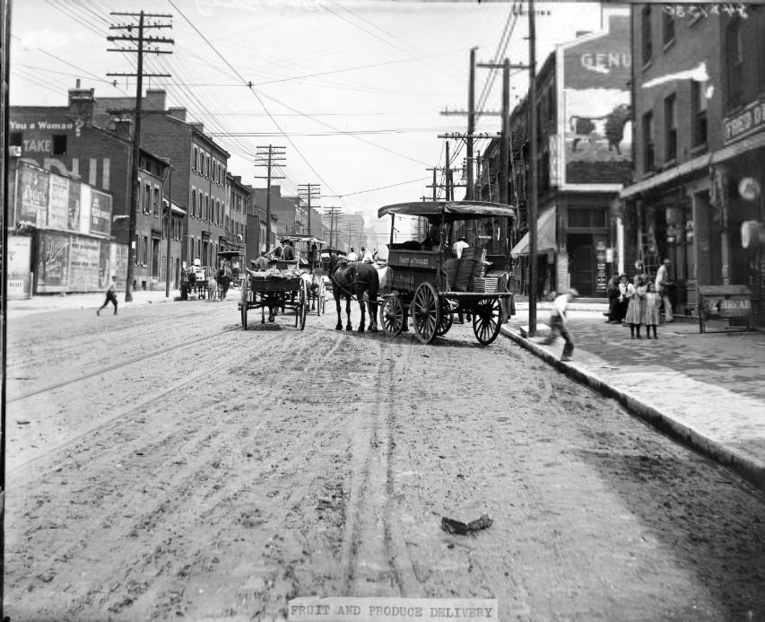 Horse-drawn wagon delivering fruit and produce to a business, 1907. Other horse-drawn wagons are visible in the street. Two girls on the sidewalk are watching the delivery.