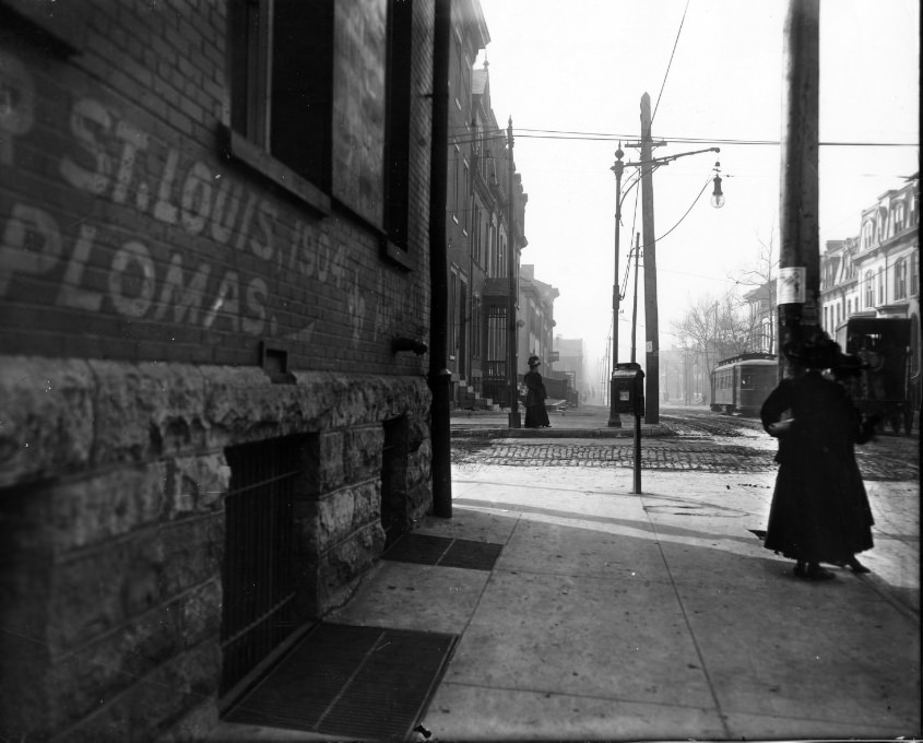 Street scene of St. Louis after during or after 1904. Two women on sidewalk. Trolley car on rails in the cobblestone street. Buildings with mansard roofs.