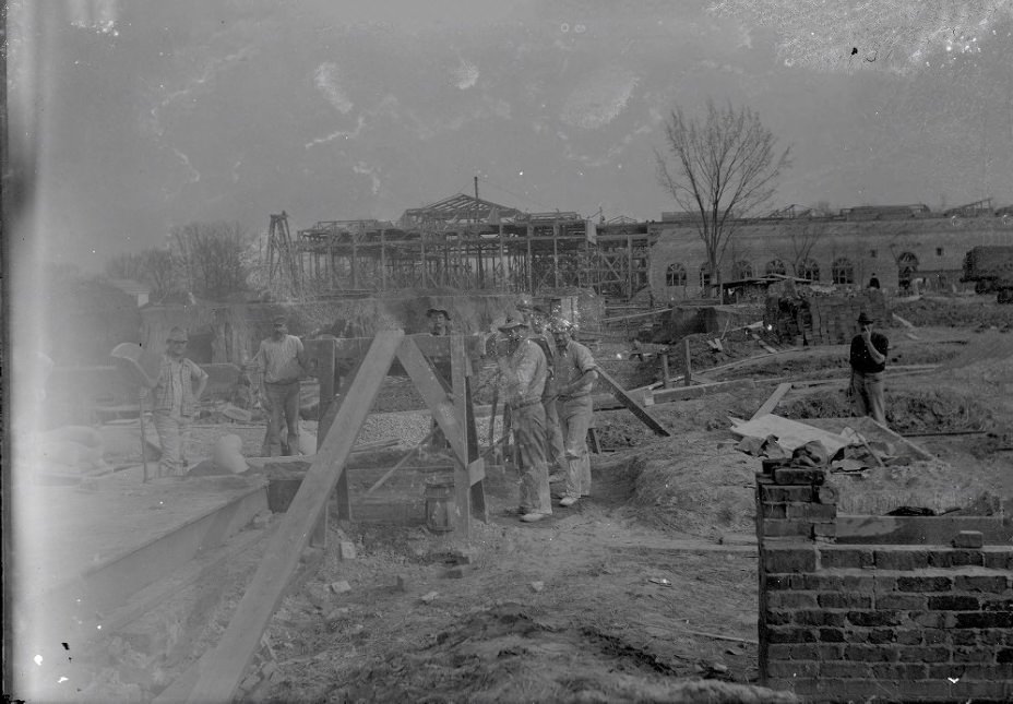 A busy construction site shows the wide skeleton of a building in the background, stacks of bricks and lumber are in the middle ground, 1907