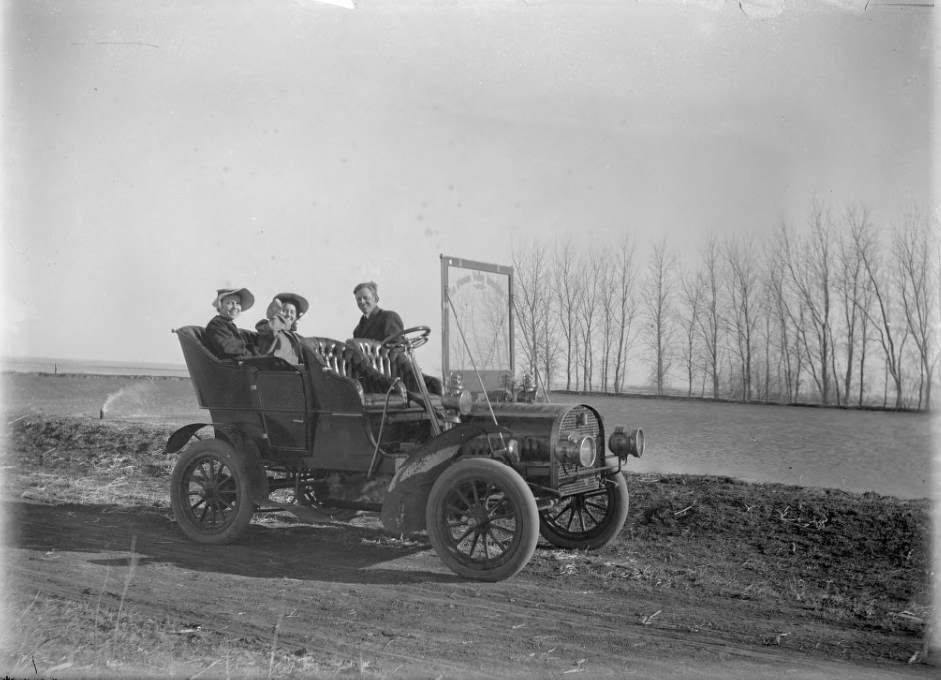 Three people in a company care parked by the river, 1909