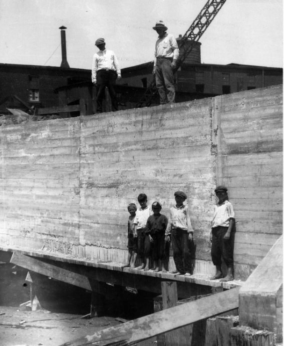 Five boys standing along the base of a poured concrete wall at a construction site, 1902