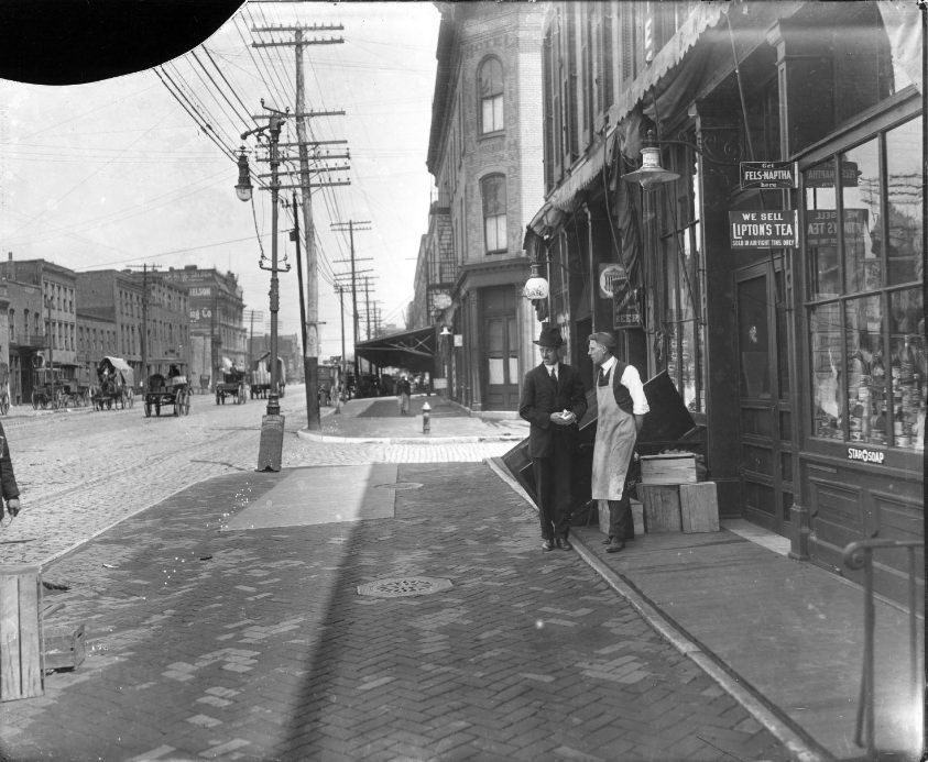 Two men standing on the sidewalk of a major street. One man appears to work at the grocery they are standing in front of, 1904