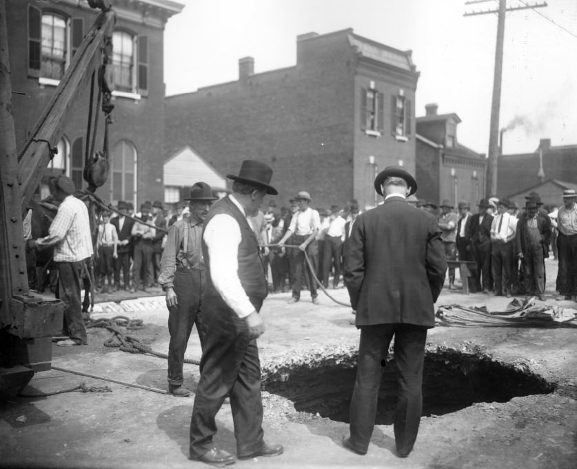 People standing around a large hole in a city street. Two men in the foreground are looking down the hole, 1903