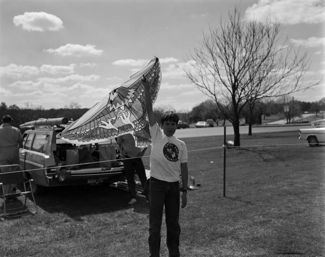 Zilker Park Kite Tournament,March 8, 1970. A child is seen holding up a kite and a vehicle is parked behind him.