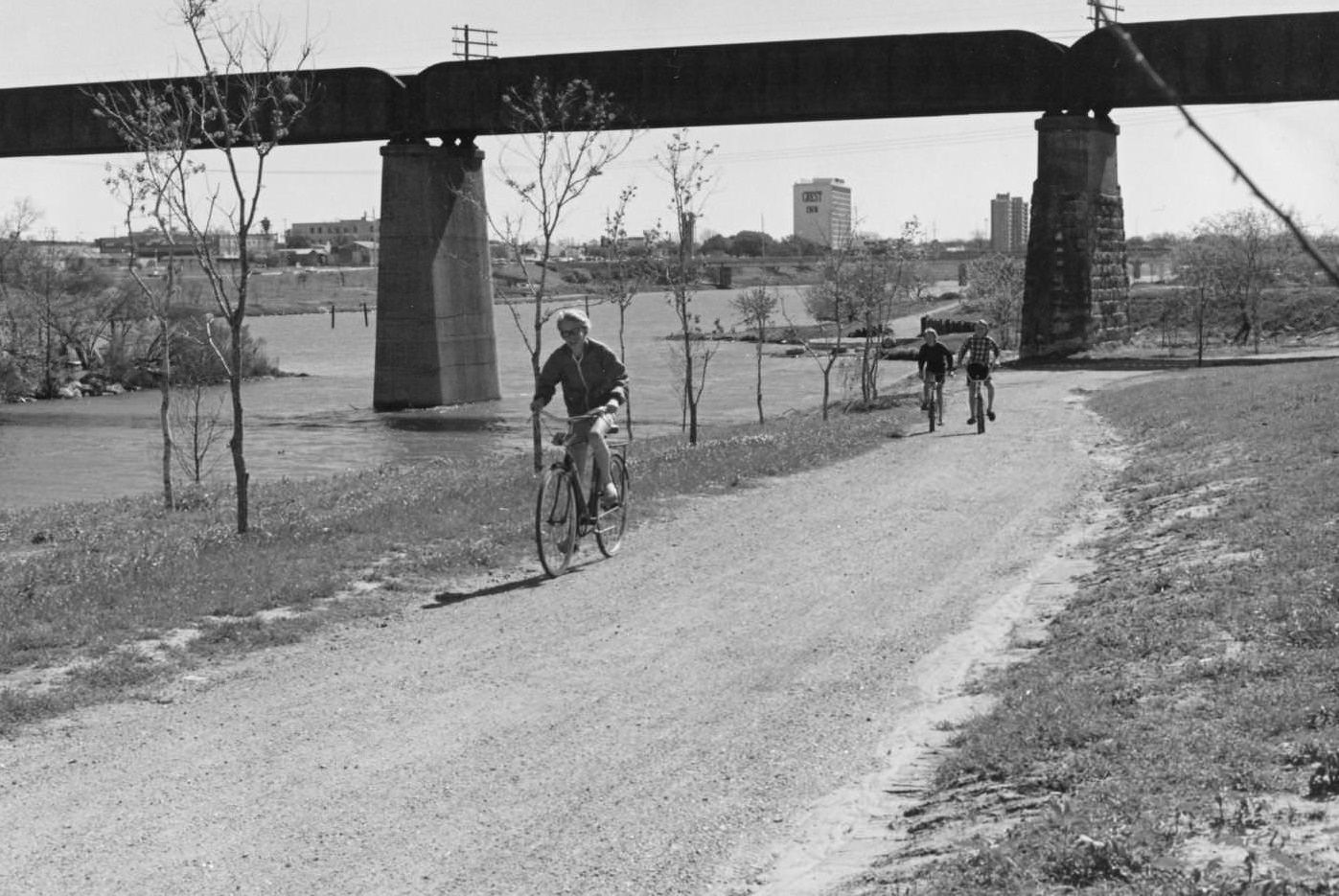 Auditorium Shores on Town Lake, looking east, with three boys riding bikes on a gravel path, 1972 .A railroad bridge crosses the river behind them. Crest Inn is visible in the distance.