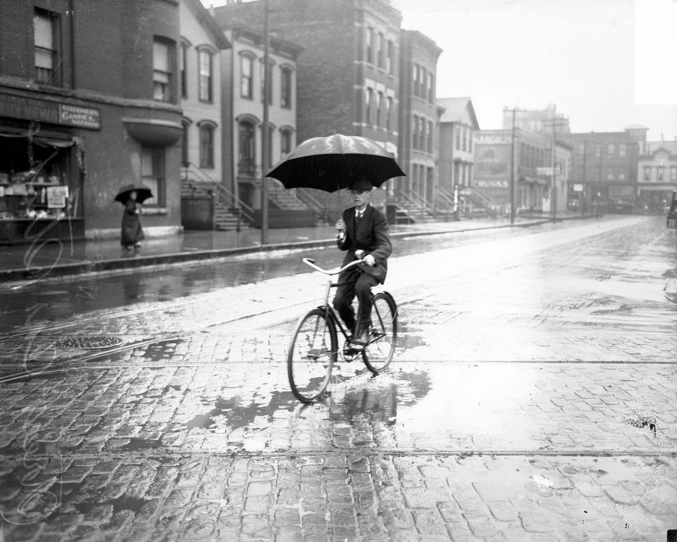 Man holding an umbrella and riding a bicycle in the rain on an empty street, with visible streetcar tracks, during a streetcar strike, Chicago, Illinois, June 15, 1915.